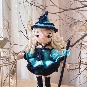 May include: A crocheted witch doll with long blonde hair, wearing a blue and black dress, a black hat with a flower, and black shoes. The doll is holding a broom.