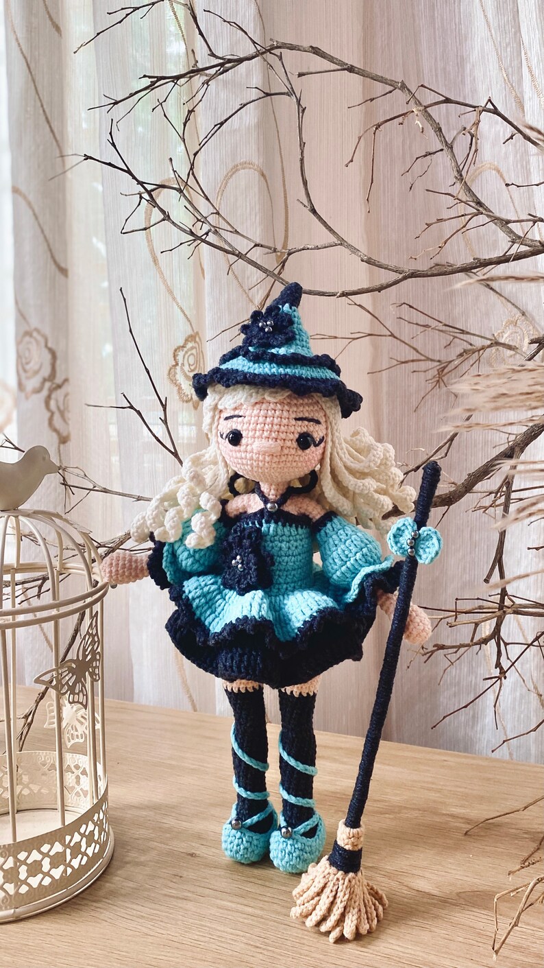 May include: A crocheted witch doll with long blonde hair, wearing a blue and black dress, a black hat with a flower, and black shoes. The doll is holding a broom.
