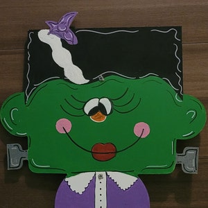 Può includere: Un mostro verde con un fiocco viola, un colletto bianco e un vestito viola. Il mostro ha i capelli neri, occhi grandi e un ampio sorriso.