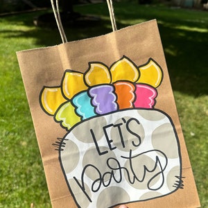 K&ouml;nnte beinhalten: Eine braune Papiert&uuml;te mit wei&szlig;em Punktmuster und dem Schriftzug "Let's Party" in Schwarz. Die Tasche hat eine wei&szlig;e Zeichnung eines Kuchens mit Regenbogenkerzen darauf.