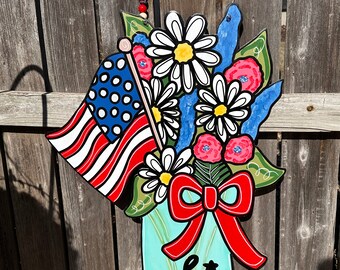 May include: A patriotic-themed door hanger featuring a mason jar with a red bow, filled with red, white, and blue flowers and a small American flag. The jar has the word "Hi" written on it.