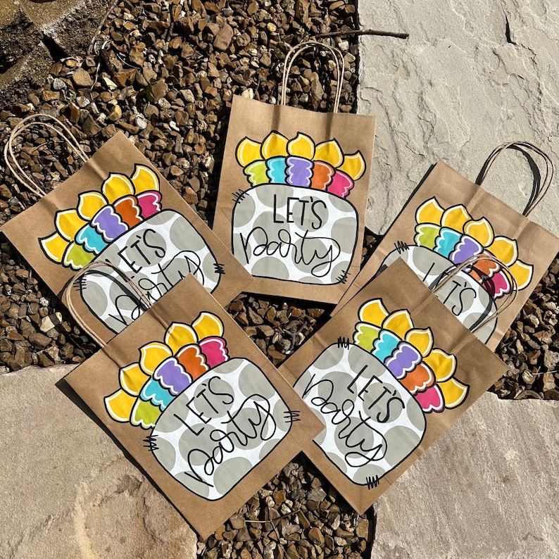 K&ouml;nnte beinhalten: Sechs braune Papiert&uuml;ten mit bunten bemalten Designs und dem Text "Let's Party" auf jeder T&uuml;te.