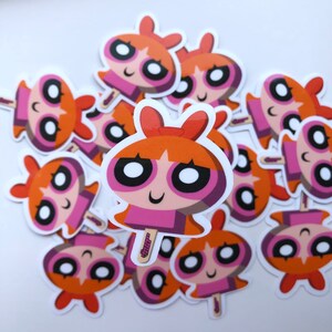 May include: A collection of stickers featuring a cartoon character with orange hair, a pink dress, and a red bow. The character has large eyes and a smiling expression. The stickers are on a white background.