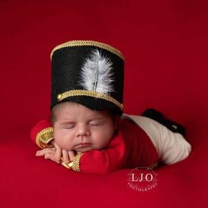 May include: A black felt hat with gold trim and a white feather plume, paired with a red long-sleeved top featuring gold cuffs and white pants. The hat is cylindrical with a flat top, and the outfit resembles a classic military uniform.