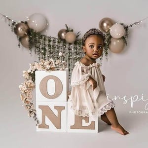 May include: A toddler wearing a beige off-the-shoulder dress with lace trim and a beaded headband, sitting beside white square blocks stacked to spell the word ONE, decorated with artificial flowers and a backdrop of balloons and greenery.