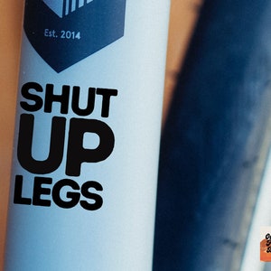 以下が含まれることがあります： 白い自転車のフレームに、「SHUT UP LEGS」と書かれた黒いステッカーが貼られています。ステッカーは自転車のフレームに、前輪の近くに貼られています。