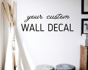 May include: A white wall with the text "your custom WALL DECAL" in black lettering. The wall is decorated with a teepee and two gray poufs.