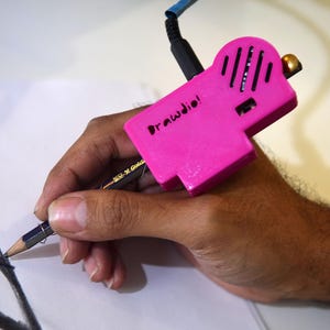 May include: A bright pink electronic device labelled Drawdio attached to a standard graphite pencil. The device features a black cable extending from the top, a speaker grille, and a small gold-coloured knob, used for drawing on white paper.