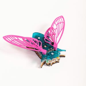 Puede incluir: Una escultura de mariposa con alas rosas brillantes y cuerpo turquesa. Las alas tienen un diseño calado. El cuerpo parece estar hecho de una placa de circuito, con pequeños componentes visibles.