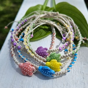 May include: A set of six colorful friendship bracelets with a turtle charm on each. The bracelets are made of white string and feature different colored beads.