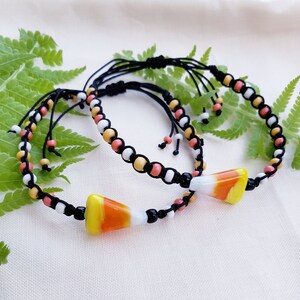 May include: Two friendship bracelets with black cord and colorful beads. Each bracelet has a candy corn charm.