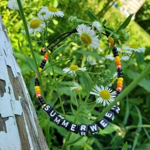 May include: A beaded bracelet with the word "SUMMERWEAR" spelled out in black letter beads. The bracelet features a pattern of black, yellow, and orange beads. The bracelet is set against a backdrop of white daisies and green foliage.