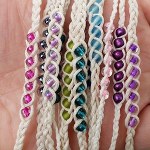 May include: A collection of handmade macrame bracelets. Each bracelet features a braided cream-colored cord with colorful glass beads in shades of pink, purple, green, and blue. The bracelets are displayed in a hand.