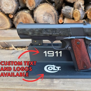 May include: A black semi-automatic pistol with a dark metal finish and textured reddish-brown wooden grips, resting on a black display stand. The stand features the white text 1911 and a Colt logo, with red arrows pointing to the text and logo area.