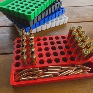 May include: A red, hexagonal-patterned ammunition tray holds several brass bullet casings and bullets. A stack of multi-colored trays, including green, black, blue, and white, sits in the background. The bullets are a copper color.