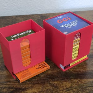 May include: Two red storage containers holding ammunition. One container has a box of Remington ammunition. The other container has a box of CCI Small Rifle Primers No. 400. The containers are on a wooden surface.