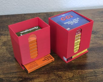 May include: Two red plastic storage dispensers for ammunition primer boxes. The left dispenser holds orange Remington boxes, while the right dispenser holds blue CCI Small Rifle Primers No. 400 boxes, with visible labels and stacked boxes inside.