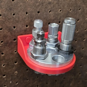 May include: A red plastic wall-mounted organizer featuring a circular aluminum base with multiple threaded holes. Several silver metal pneumatic fittings and adapters are inserted into the base, with one empty threaded port visible.