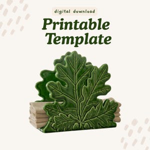 May include: A green ceramic napkin holder shaped like an oak leaf with two small acorns at the base. The text digital download Printable Template is displayed above the object. The holder contains a stack of folded beige napkins.