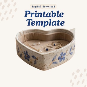 May include: A heart-shaped ceramic jewelry dish with a speckled cream finish and hand-painted blue floral motifs. The interior is lined with beige fabric and contains a gold chain necklace, two rings, and two stud earrings. Text reads: digital download Printable Template.