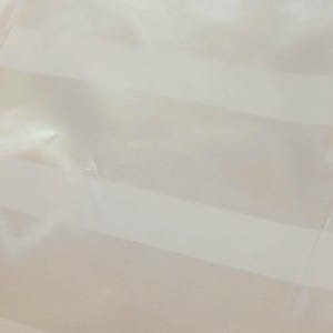 May include: Close-up of a sheer, cream-colored fabric with horizontal stripes. The fabric appears to be lightweight and slightly translucent, with a subtle sheen. The stripes are a lighter shade of cream.