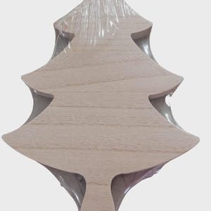 May include: A light-colored wooden serving board shaped like a Christmas tree with a smooth, natural grain finish. The board features a small circular hole at the base of the trunk and is encased in a clear, tight-fitting plastic shrink wrap.