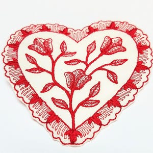 May include: A heart-shaped fabric patch featuring an off-white base with red embroidered floral motifs in the center. The edge is finished with a decorative red scalloped lace trim that follows the heart shape of the patch.