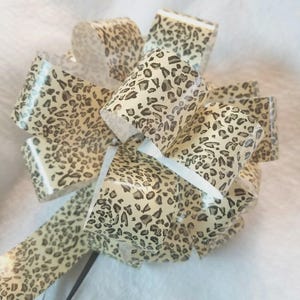 May include: A decorative bow made of tan and brown leopard print ribbon. The bow is tightly wound with multiple loops and tails, perfect for gift wrapping or crafting. The ribbon has a glossy finish.