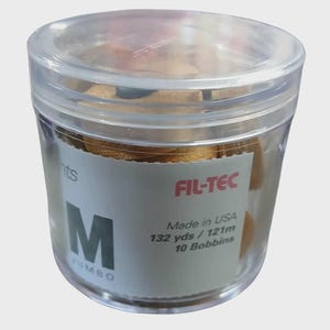 May include: A clear plastic container with a lid, filled with gold-colored thread bobbins. The label reads "FIL-TEC" and "Made in USA, 132 yds / 121m, 10 Bobbins". The container is labeled "M JUMBO".