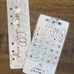 May include: Two clear plastic cards. One is a bookmark with the words "NO SPEND CHALLENGE". The other is a calendar with numbered boxes for a one-month "NO SPEND CHALLENGE". The cards are designed to track spending.