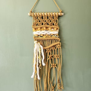 Puede incluir: Tapiz de macramé hecho a mano en tonos amarillo mostaza, crema y madera natural. La pieza tejida presenta intrincados patrones de nudos, cuentas de madera y largas borlas. Una pieza decorativa para decoración bohemia o moderna.