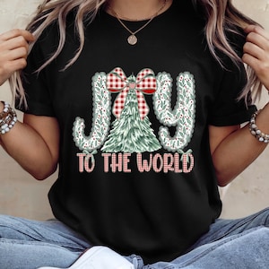 May include: Black t-shirt with a Christmas-themed graphic. The design features the words "JOY TO THE WORLD" in a festive font, with a Christmas tree and a red and white plaid bow. The letters are decorated with green leaves.