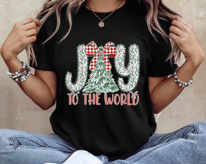 May include: Black t-shirt with a Christmas-themed graphic. The design features the words "JOY TO THE WORLD" in a festive font, with a Christmas tree and a red and white plaid bow. The letters are decorated with green leaves.
