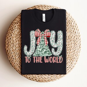 May include: Black t-shirt with the words "JOY TO THE WORLD" in a festive design. The word "JOY" is in a decorative font with a green and white pattern. A Christmas tree and a red and white plaid bow are in the center.