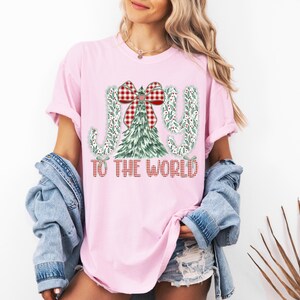 May include: Pink t-shirt with a Christmas-themed graphic. The design features the words "JOY TO THE WORLD" in a plaid pattern, with a Christmas tree and bow. The shirt is a light pink color.