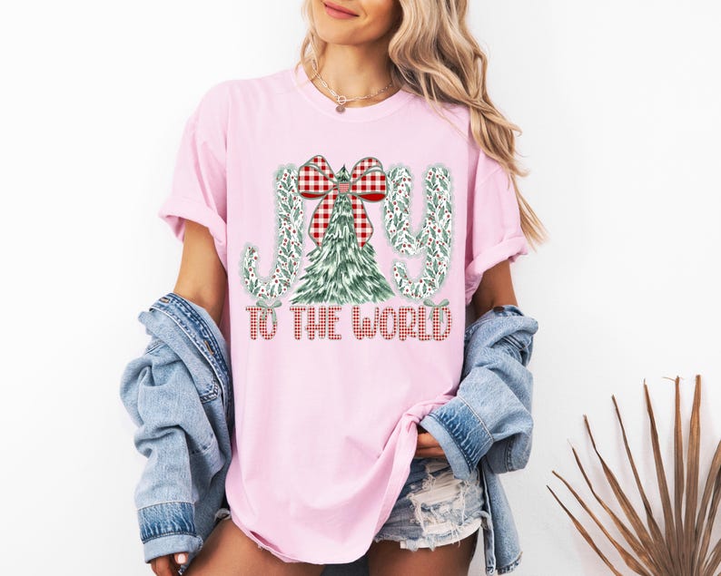 May include: Pink t-shirt with a Christmas-themed graphic. The design features the words "JOY TO THE WORLD" in a plaid pattern, with a Christmas tree and bow. The shirt is a light pink color.