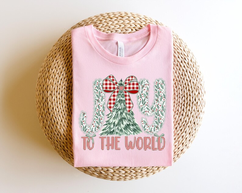May include: A light pink t-shirt with a Christmas-themed graphic. The graphic includes a green Christmas tree, a red and white plaid bow, and the words "JOY TO THE WORLD" in a festive font. The shirt is laid on a woven, circular surface.