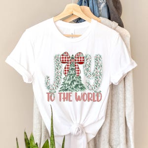 May include: White t-shirt with a Christmas-themed design. The design features the words "JOY TO THE WORLD" in red, with the letters "J" and "Y" decorated with green leaves and berries. A Christmas tree and a red and white plaid bow complete the design.