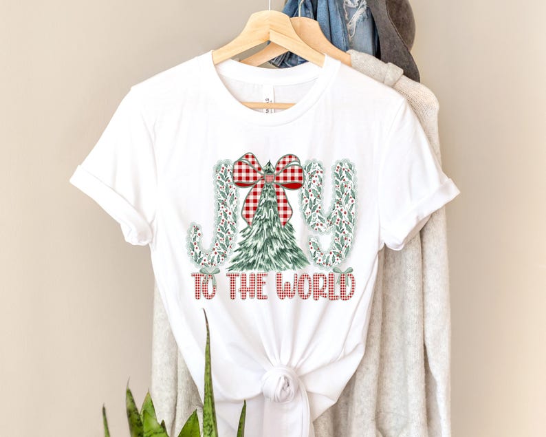 May include: White t-shirt with a Christmas-themed design. The design features the words "JOY TO THE WORLD" in red, with the letters "J" and "Y" decorated with green leaves and berries. A Christmas tree and a red and white plaid bow complete the design.