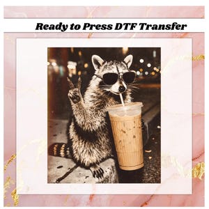 May include: DTF transfer featuring a raccoon wearing sunglasses, giving a peace sign, and drinking iced coffee. The raccoon is in a city setting. The text "Ready to Press DTF Transfer" is at the top.