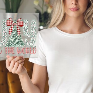 May include: A clear, rectangular sign with the words "JOY TO THE WORLD" in red and white checkered font. The design includes a Christmas tree and bow, with green and white accents. The sign is held up by a person.