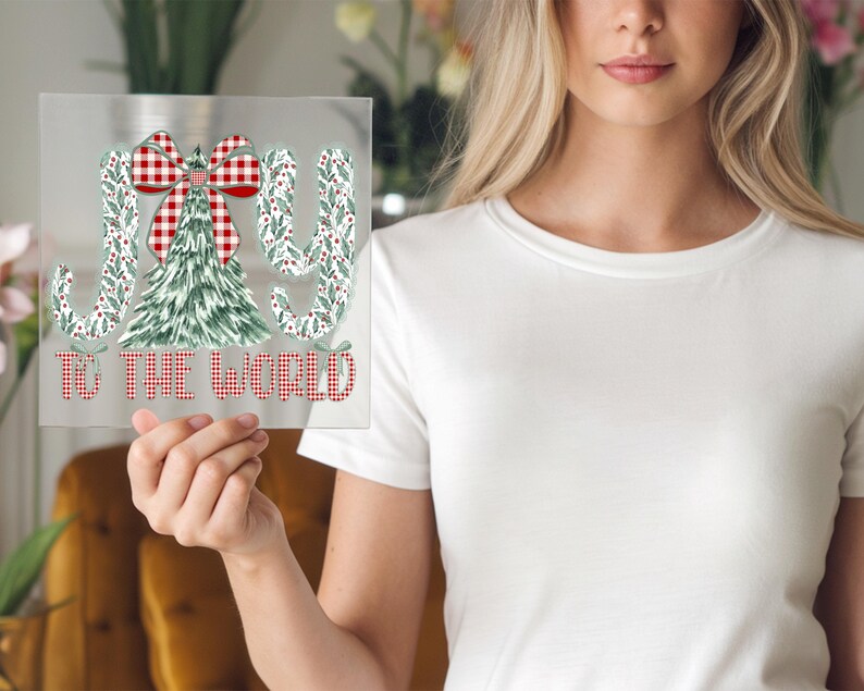 May include: A clear, rectangular sign with the words "JOY TO THE WORLD" in red and white checkered font. The design includes a Christmas tree and bow, with green and white accents. The sign is held up by a person.