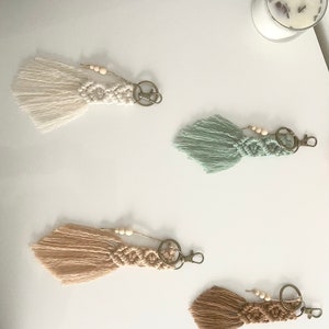 May include: Four handmade macrame keychains with wooden beads and a metal ring. The keychains are in the colors white, light green, beige, and brown.
