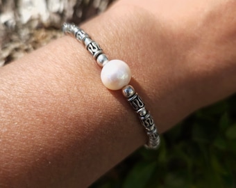 May include: A silver bracelet with a single white pearl charm. The bracelet is made of small, round beads with a decorative design.