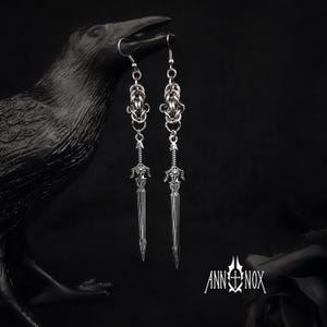 May include: Silver-toned earrings featuring a sword design. The earrings have a chainmail-style top and a detailed sword pendant. The image is set against a dark background with a raven sculpture and the text "ANN+NOX".