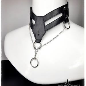 May include: Black leather choker necklace with a silver chain and ring accents. The choker features a geometric design with a cross pendant and is displayed on a white mannequin. The Ann Nox logo is visible in the bottom right corner.