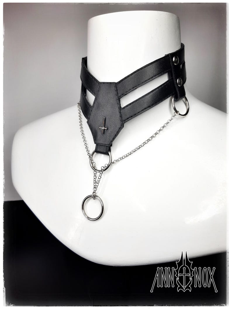 May include: Black leather choker necklace with a silver chain and ring accents. The choker features a geometric design with a cross pendant and is displayed on a white mannequin. The Ann Nox logo is visible in the bottom right corner.