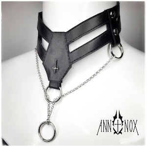 May include: Black faux leather choker necklace with a coffin-shaped centerpiece and a silver inverted cross. The choker features silver chain detailing and a large silver ring. The brand name "ANN NOX" is printed in black.