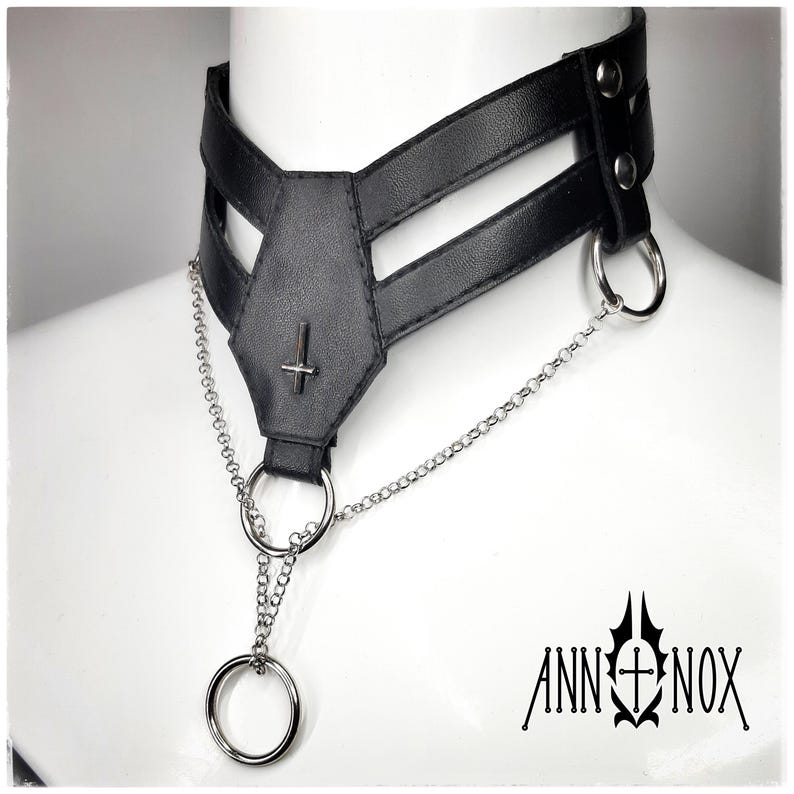 May include: Black faux leather choker necklace with a coffin-shaped centerpiece and a silver inverted cross. The choker features silver chain detailing and a large silver ring. The brand name "ANN NOX" is printed in black.