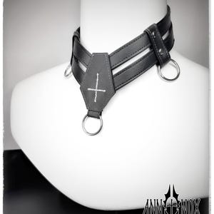 May include: A black leather choker with a cross pendant and silver metal rings. The choker has a layered design and is displayed on a white mannequin head. The brand name "ANN INOX" is visible at the bottom.